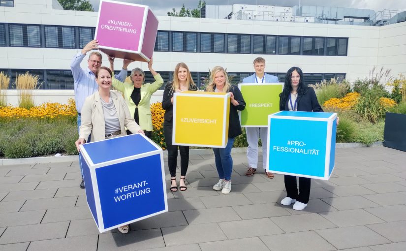 Unternehmenswerte kreativ präsentiert: Birgit Aspin (Chief People Officer und Mitglied der Geschäftsleitung) sowie (v. l.) Frank Schultz (Abteilungsleiter Direktservice und Ausbildungswerkstatt), Nadja Bürger (Leiterin Geschäftsfeld Ambulantes Gesundheitswesen, Direktservice und Ausbildungswerkstatt und Mitglied der Geschäftsleitung) und Leonie Münnich (Direktservice-Team / Ausbildungswerkstatt), Pia Stohlmann (Referentin Personalentwicklung) begrüßen mit Niklas Kehne (Auszubildender zum Kaufmann für Versicherungen und Finanzen) und Lilli Richter (Auszubildende zur Kauffrau für Versicherungen und Finanzen) zwei der neuen Auszubildenden. (Foto: Ecclesia Gruppe)